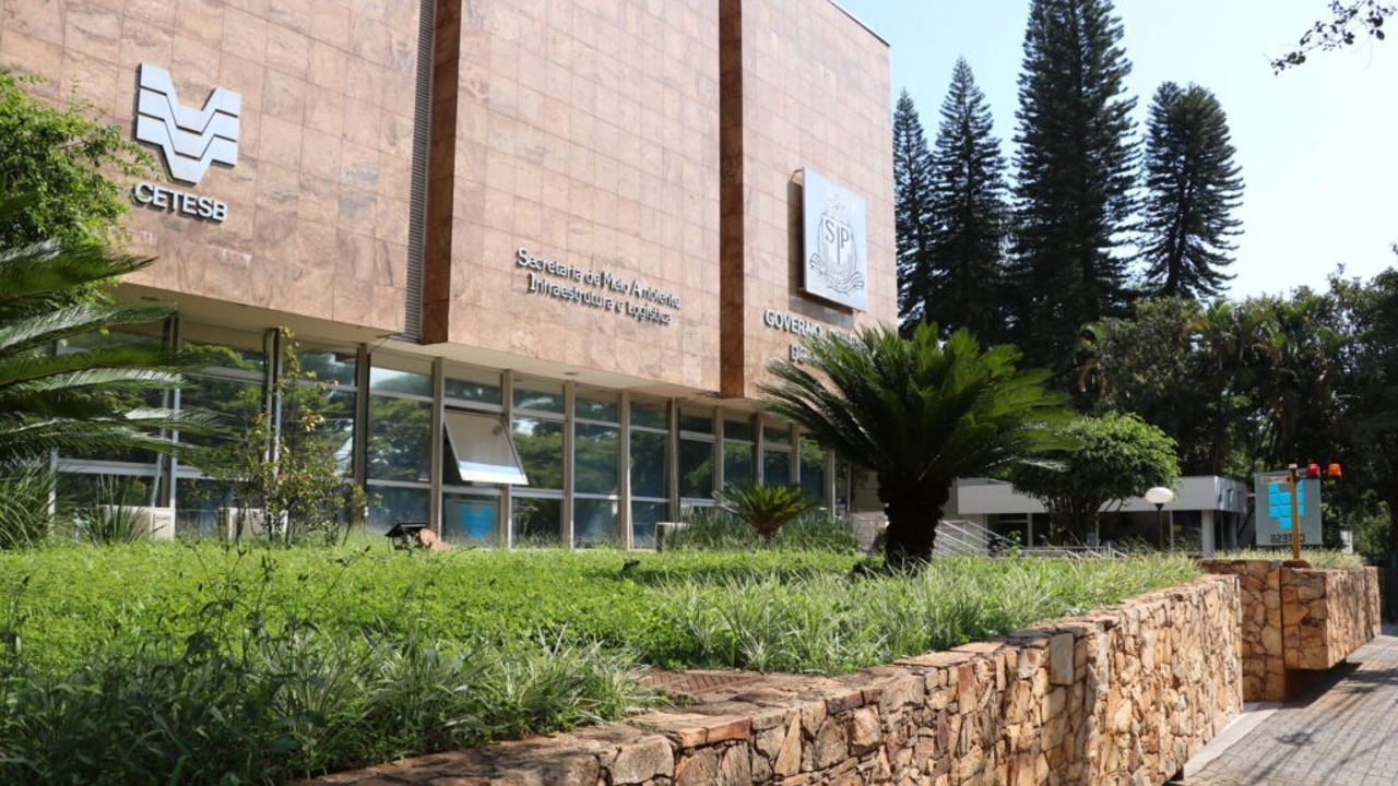 Fachada do prédio da sede da CETESB com jardim e árvores ao redor em dia ensolarado.